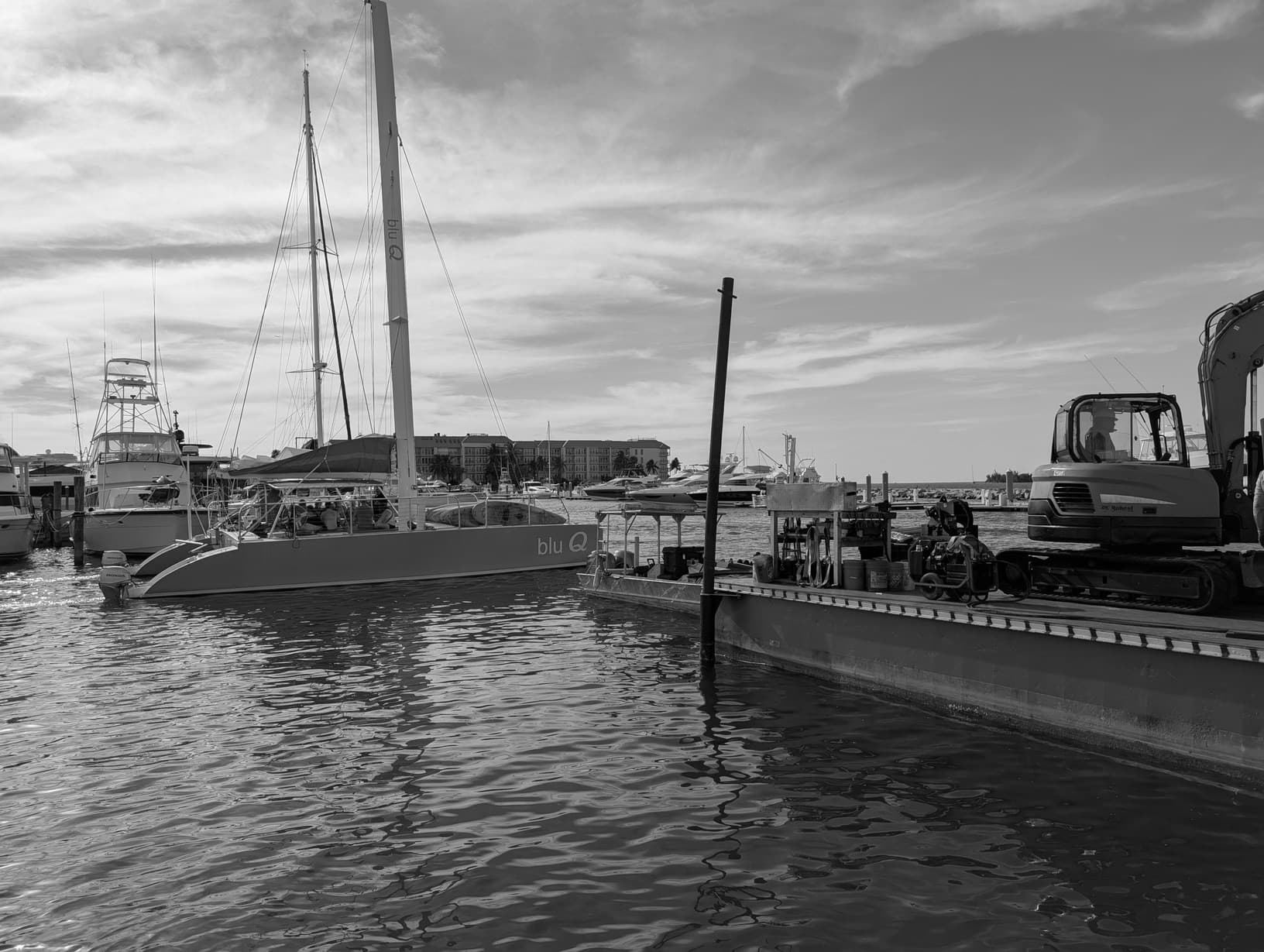 Work barge with an excavator moored beside a sailing catamaran in a marina
